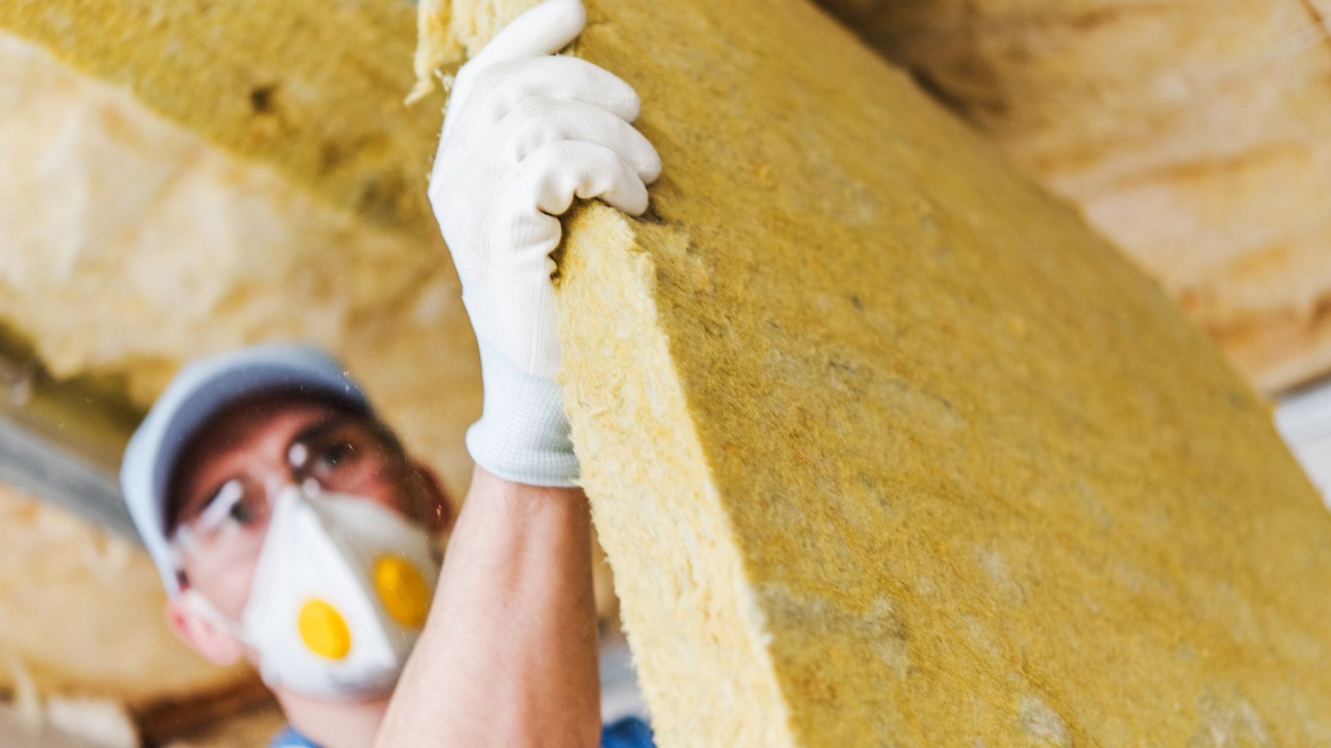 A worker in safety gear, including a mask and gloves, lifts and installs a large piece of yellow fiberglass insulation in an attic or ceiling space.