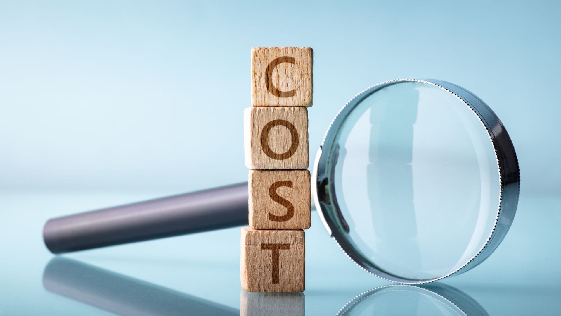 Three wooden blocks stacked vertically, spelling the word "COST," with a magnifying glass beside them, symbolizing financial analysis or cost estimation.