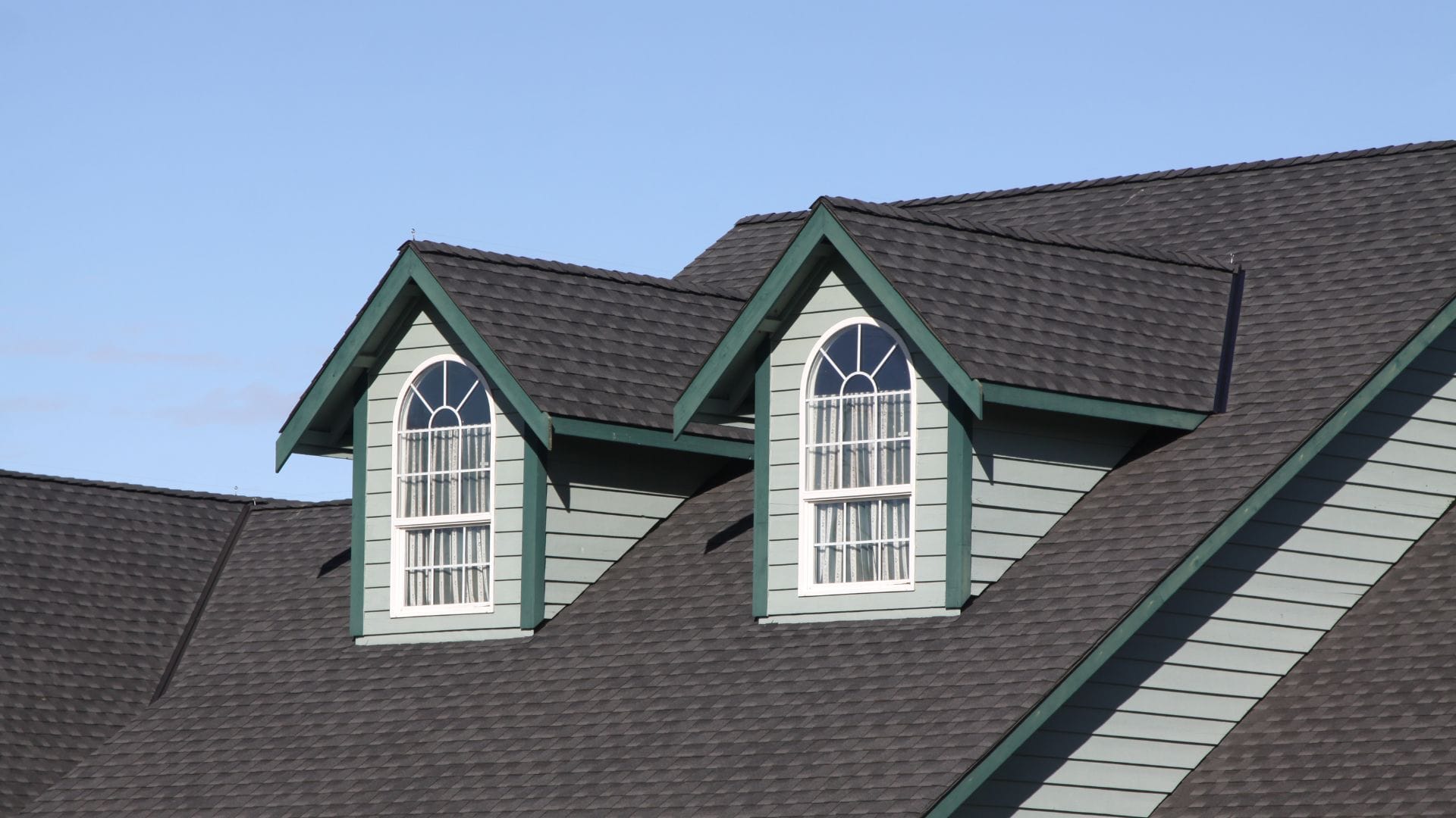 A residential home with dark grey asphalt shingles, featuring two dormer windows with green trim and decorative arches.
