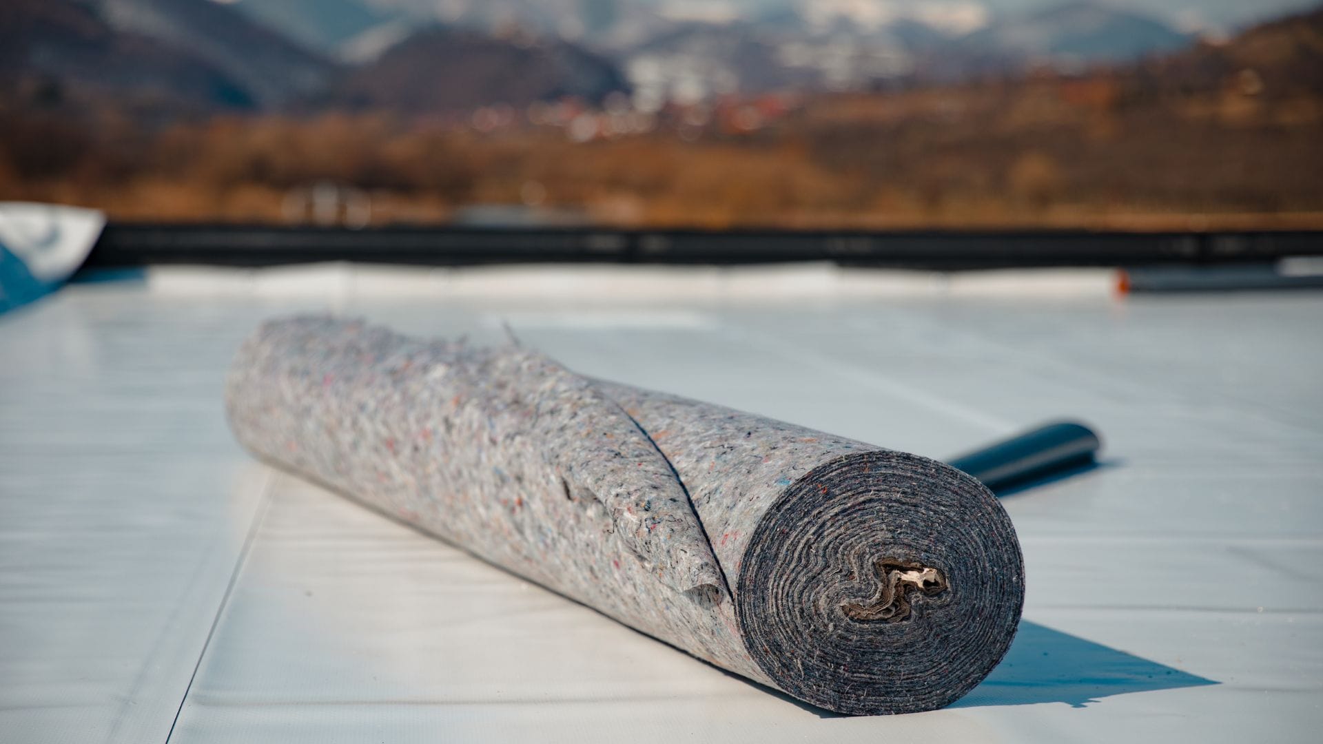 A roll of roofing underlayment on a flat rooftop, with a scenic mountain landscape in the background.