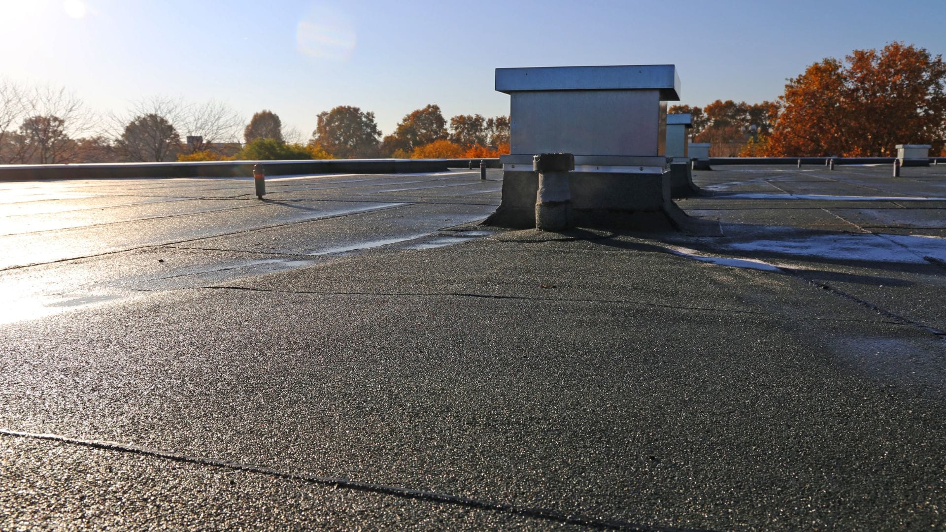 Close-up view of a flat roof with a ventilation unit and sunlight glinting off the waterproof surface.