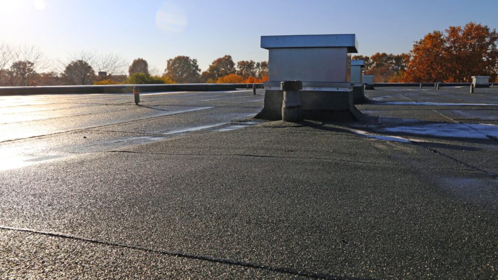 Flat commercial roof with small puddles of water and vent structures under clear sky.