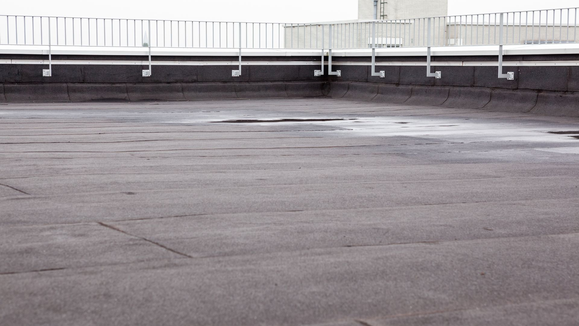 Flat commercial rooftop covered with a dark membrane surface, surrounded by a white metal safety railing and showing slight water pooling, with urban buildings in the background.
