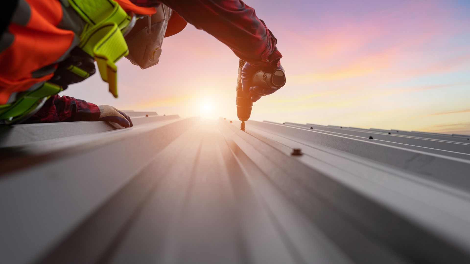 Worker wearing safety gear installs a corrugated metal roof panel with a power tool at sunset, highlighting the metallic texture under the colourful sky.