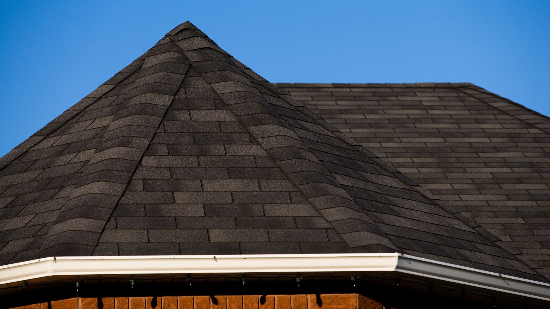 Close-up view of a pitched roof with brown metal roof tiles and multiple intersecting ridges, set against a cloudy sky and forest background.